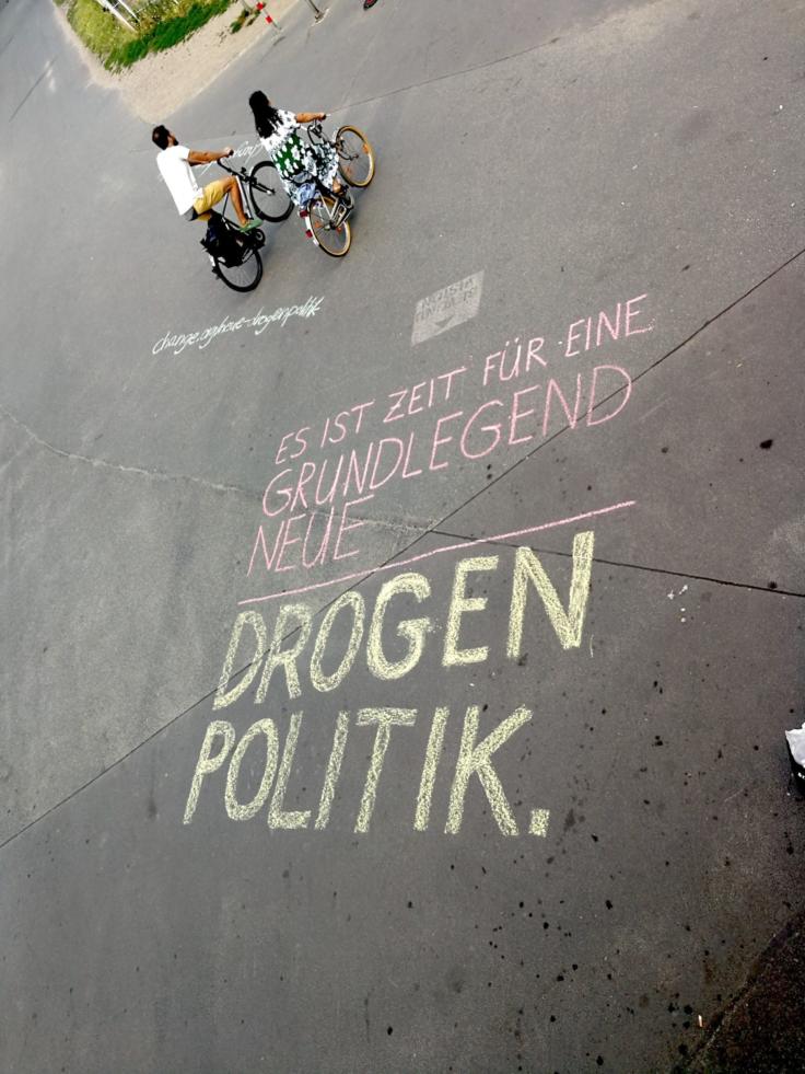 "Es ist Zeit für eine grundlegend neue Drogenpolitik" mit Straßenmalkreide auf die Straße gemalt und zwei Radfahrer